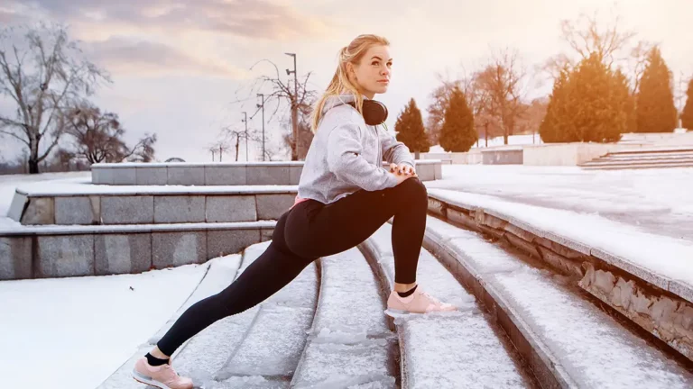 Žena na zimních schodech, provádějící dynamický warm up, oblečená v zimním běžeckém outfitu s kompresními podkolenkami, mikinou a bundou, připravující se na trénink.“